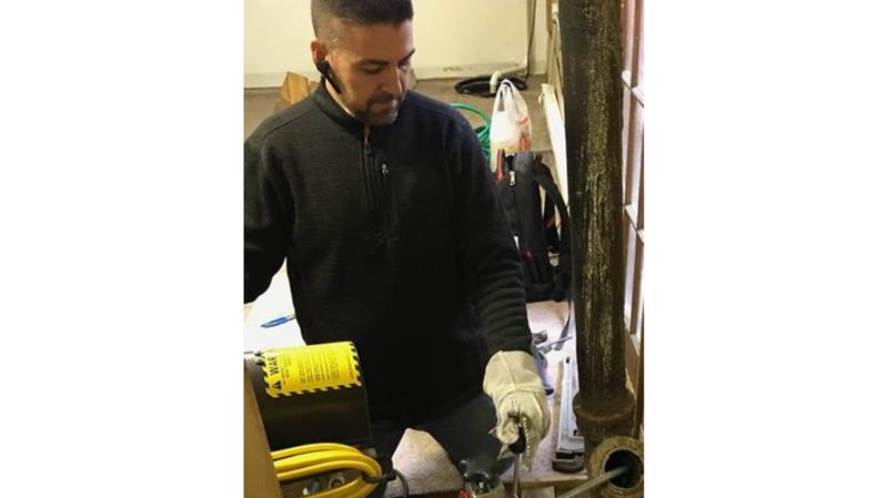 A man wearing gloves and a headset uses a tool to work on machinery indoors, with cables and equipment visible around him.