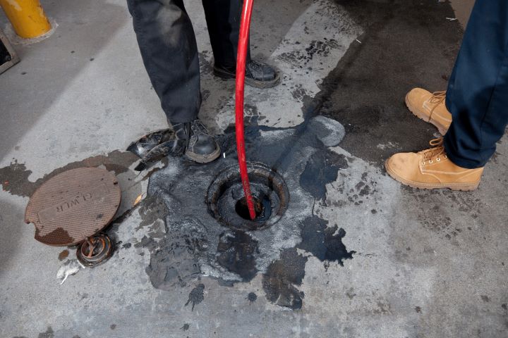 Two people stand by an open manhole on a concrete surface, one holding a red hose inserted into the hole. Black liquid stains surround the opening, and a removed metal cover lies nearby.