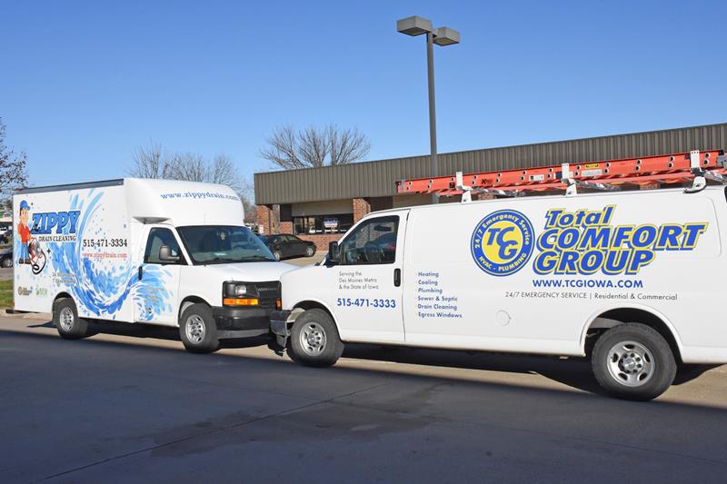 Two white service vans are parked side by side in a lot. One van advertises "Zippy Water Solutions," and the other advertises "Total Comfort Group" with HVAC and plumbing services. A building is visible in the background.