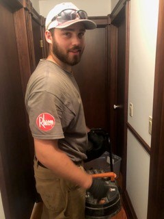 A man wearing a cap, work gloves, and a Rheem-branded shirt stands in a narrow hallway, holding a bucket with a handle and looking back at the camera.