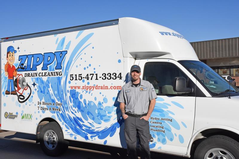 A man in a gray uniform stands in front of a white Zippy Drain Cleaning van with blue swirl graphics and company contact information, parked outside a building on a sunny day.