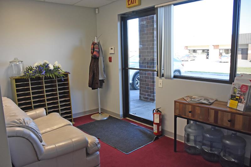 Small office lobby with a light leather couch, mail slots, coat rack with jackets, fire extinguisher, water dispensers, a side table with pamphlets, and a glass door with a window showing cars and buildings outside.
